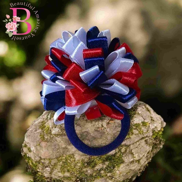 Red, white and navy blue ribbon pom pom hair tie displayed on a mossy rock outdoors with soft natural background lighting.