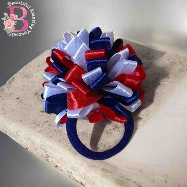 Red, white and navy blue ribbon pom pom hair tie on a navy elastic band, displayed on a stone surface with soft lighting.