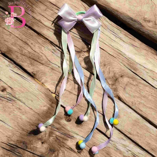 Pastel satin bow hair tie with flowing ribbon tails and colorful pom pom accents displayed on rustic wooden background outdoors.