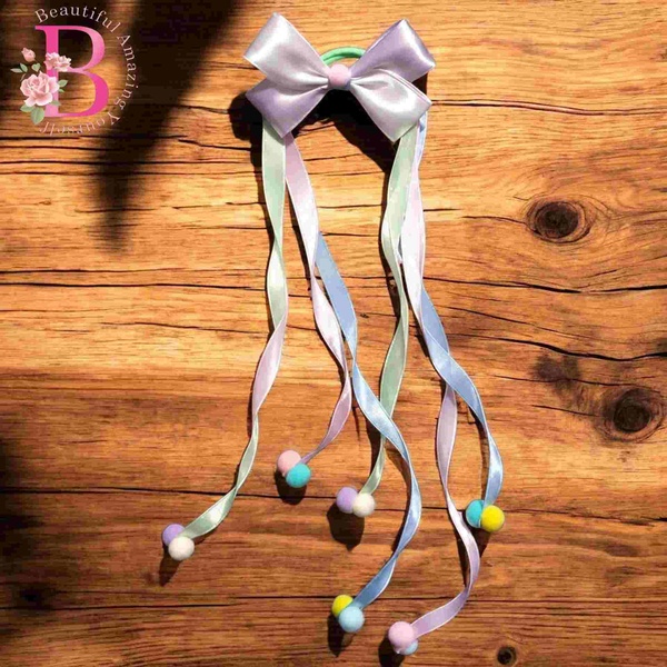 Pastel satin bow hair tie with long ribbon streamers and colorful pom pom accents displayed on a rustic wooden background.