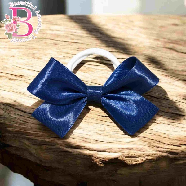 Navy blue satin hair bow on white elastic band displayed on rustic wood surface with soft sunlight and elegant boutique styling.