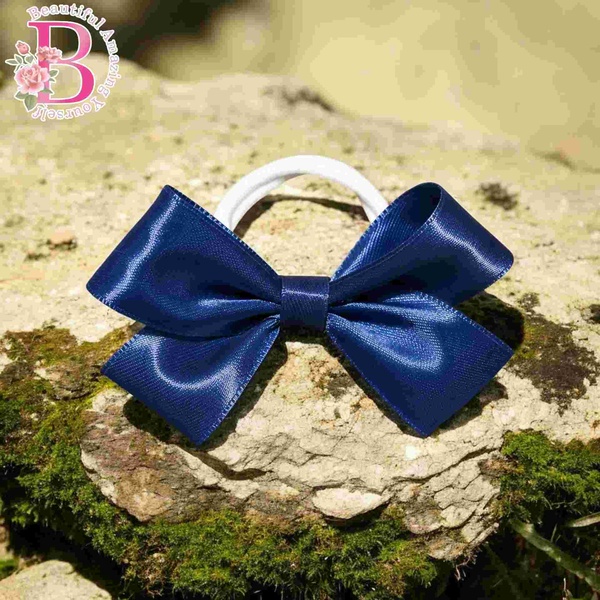 Navy blue satin ribbon hair bow on a white elastic band displayed on mossy rocks outdoors with a glossy finish and neat loops.