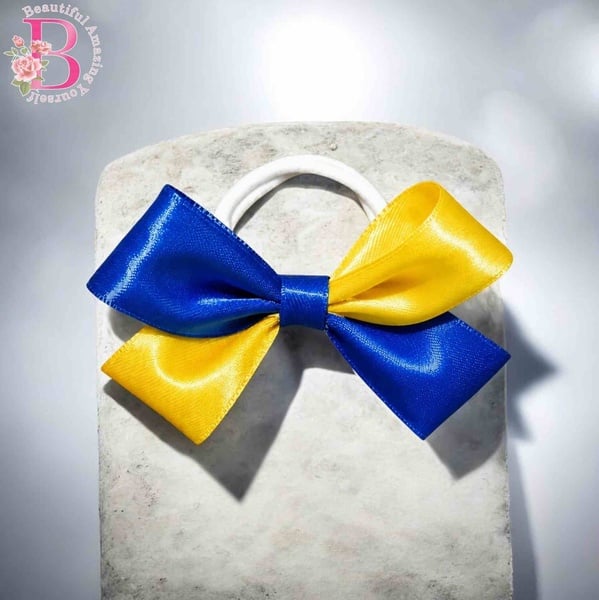 Royal blue and canary yellow satin hair bow on white elastic, displayed on a stone block with soft lighting and logo top left detail..