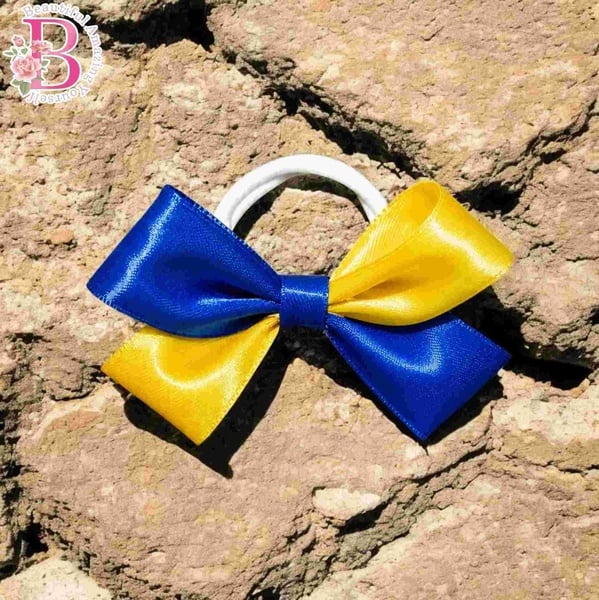 Royal blue and canary yellow satin hair bow on a white elastic band displayed on textured rock with glossy ribbon finish outdoors.