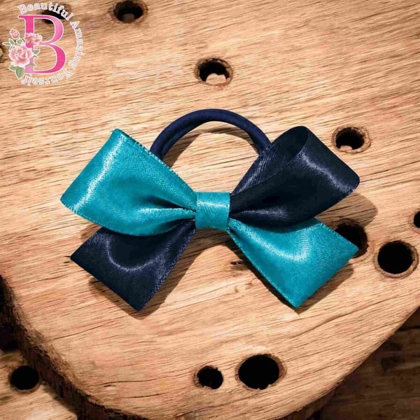 Turquoise and navy blue satin hair bow on a navy elastic band displayed on a rustic wooden surface with decorative drilled holes.
