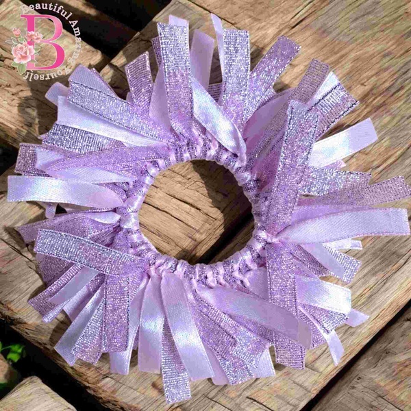 Light pink glitter ribbon scrunchie with satin and shimmer strands arranged in a circular design on a rustic wooden surface outdoors.