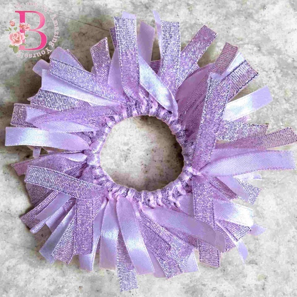 Light pink glitter ribbon scrunchie with shimmering satin strands arranged in a circular style on a textured stone background.