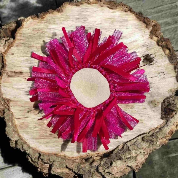 Bright pink ribbon scrunchie hair accessory on a natural wood slice background with layered textured ribbons and elastic center detail.