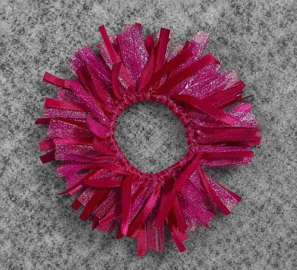 Pink metallic ribbon scrunchie with layered textured strands on a grey granite background, viewed from above in soft light.