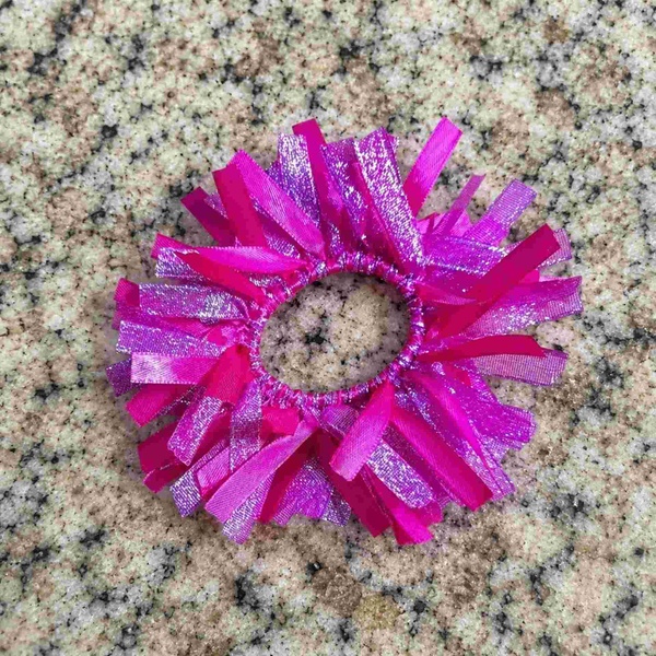 Bright pink ribbon hair scrunchie with glittery streamers, centered on a speckled beige and gray granite background in view.