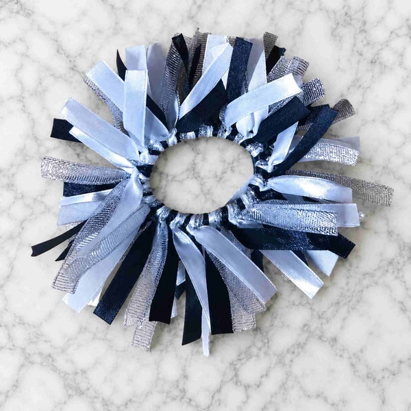 Black, white and silver ribbon scrunchie on a white marble background, featuring layered satin and metallic ribbon strands in a circular design.