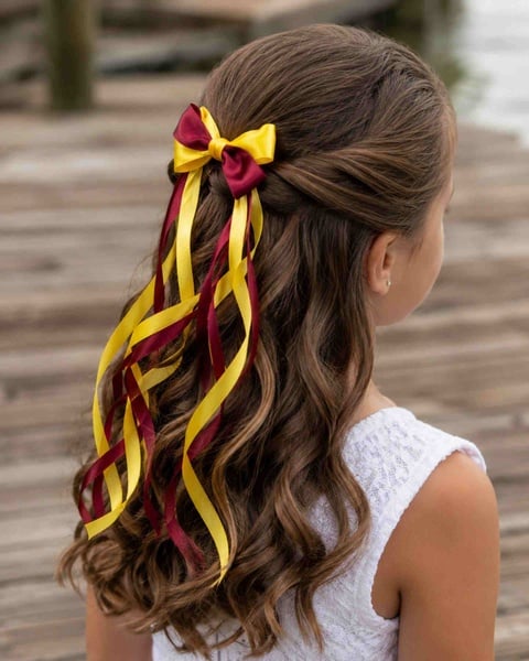 Girl viewed from behind at an angle wearing a maroon and yellow twirl ribbon bow with flowing curls in long brunette hair.