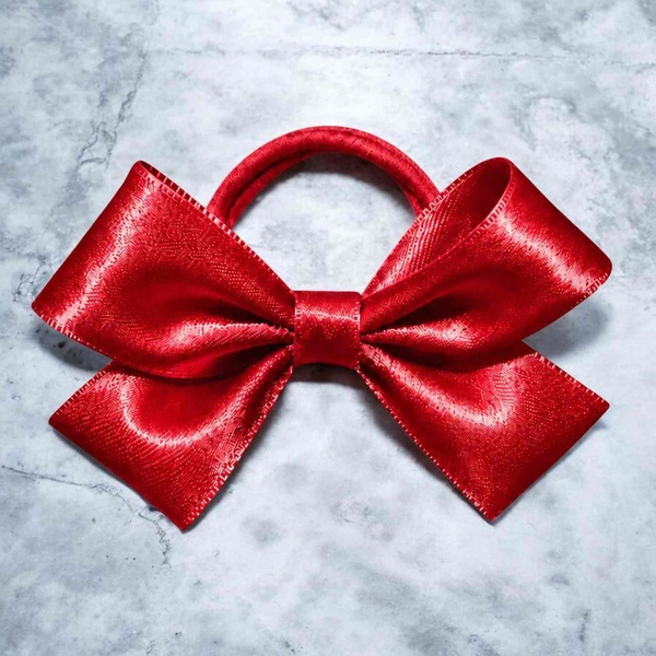 Shiny red satin hair bow with wide loops and tails attached to a matching elastic band, displayed on a light gray marble surface.