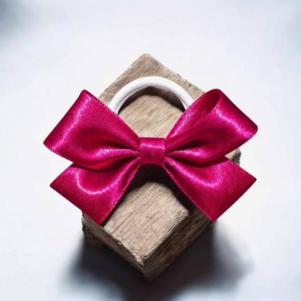 Bright fuchsia satin ribbon bow attached to a white elastic hair tie, displayed on a rustic wooden block against a soft light background.