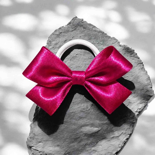 Bright fuchsia satin ribbon bow on a white elastic hair tie, displayed on a gray stone surface with glossy sheen and elegant folds.