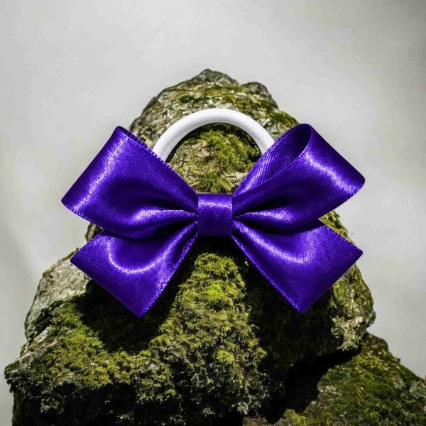 Purple satin ribbon bow on a white elastic hair tie displayed on a moss-covered rock, featuring glossy loops and a matching center wrap detail.