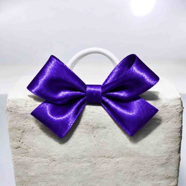 Purple satin hair bow on a white elastic band, displayed on a textured stone block with glossy ribbon loops and a matching center wrap.