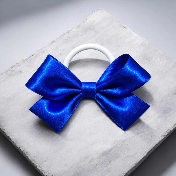 Blue satin hair bow with a white elastic band, featuring glossy ribbon loops and tails, displayed on a light textured square backdrop.