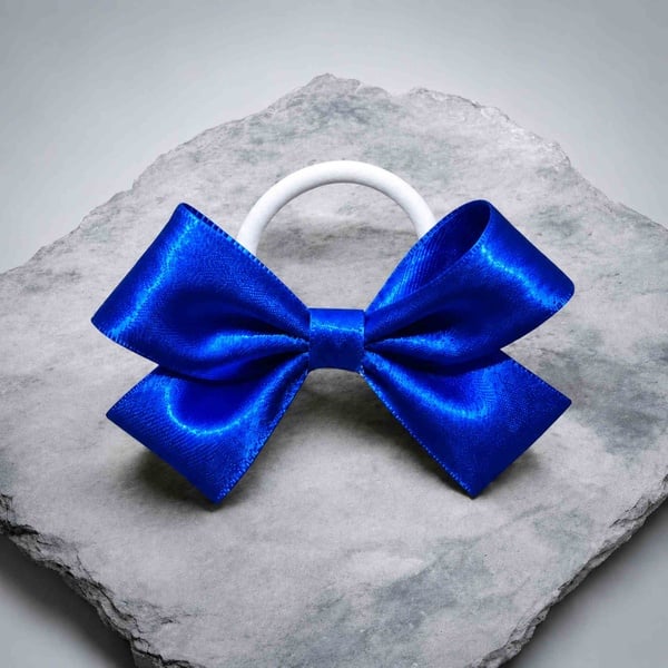 Royal blue satin hair bow with a white elastic band, displayed on a textured stone surface, featuring glossy loops and elegant styling.