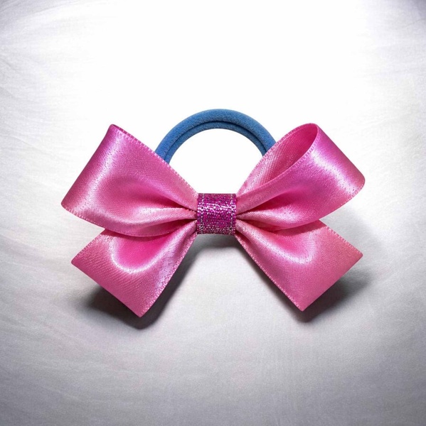 Pink satin ribbon bow hair tie with a glittery center band and blue elastic loop, displayed on a light background with a glossy finish.