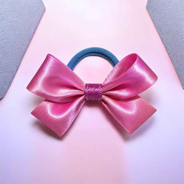 Pink satin ribbon hair bow with glitter center on a blue elastic band, displayed on a soft pink background with a glossy finish.