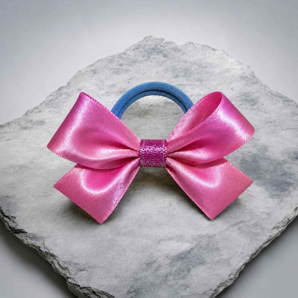 Pink satin ribbon bow hair tie with glitter center and blue elastic band, displayed on a textured gray stone slab background.
