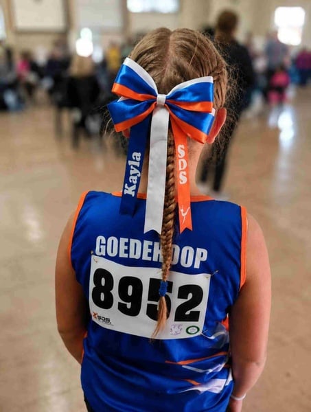 Girl viewed from behind in a blue and orange athletics uniform with a personalized ribbon bow and braid at an indoor event.