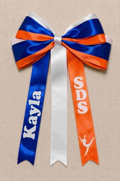 Royal blue, orange, and white layered cheer bow with custom names and SDS on ribbon tails, displayed on neutral linen fabric.