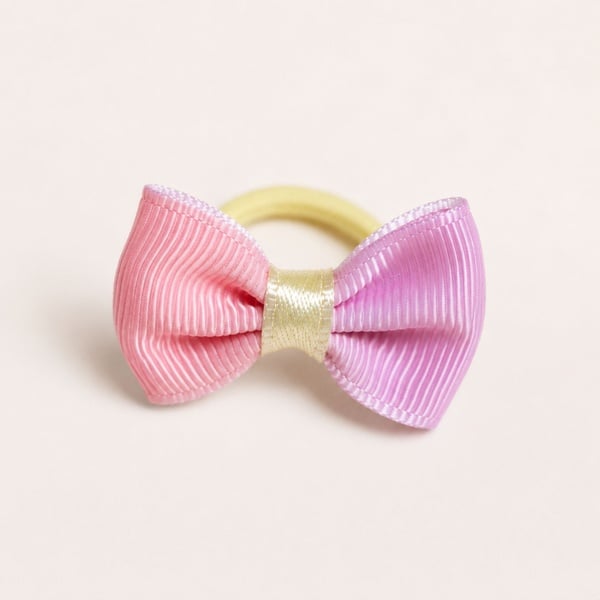 Pink and lilac grosgrain ribbon hair bow with a gold satin centre wrap on a soft cream background, attached to a yellow hair tie.