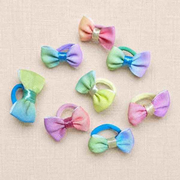 Pastel grosgrain ribbon bow hair ties in soft rainbow colours arranged on a light beige linen background, ideal for girls' hair accessories.