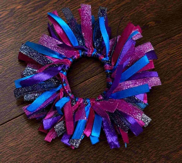 Pink, purple, blue, and black glitter ribbon scrunchie with layered satin and shimmer strands arranged in a circular design on a dark wood surface.