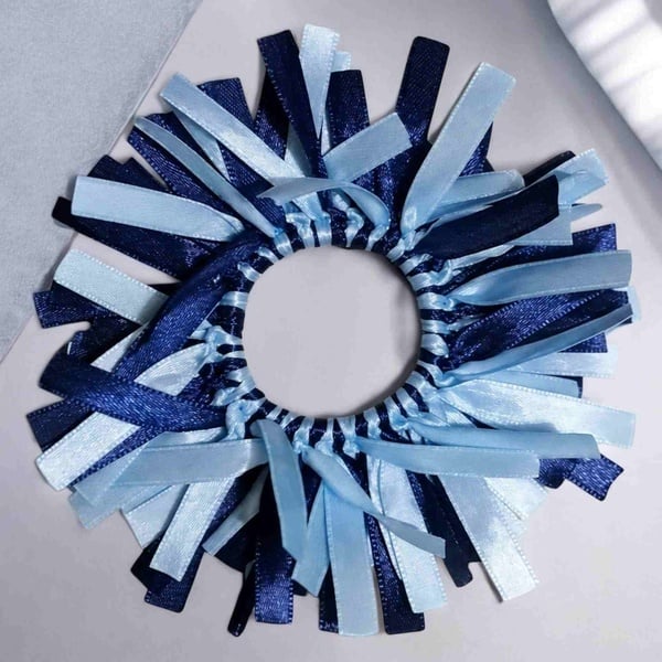 Blue and light blue ribbon scrunchie with layered satin strands, arranged in a circular design on a soft neutral background.