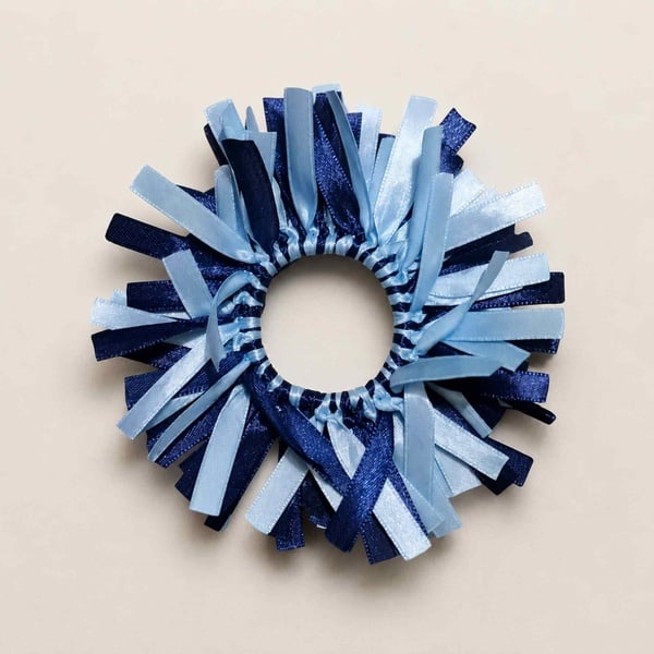 Navy and light blue ribbon scrunchie featuring layered satin streamers, displayed in a circular shape on a soft beige background.