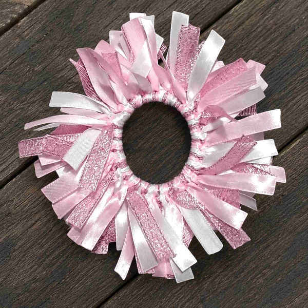 Pink and white ribbon scrunchie with satin and glitter strands arranged in a fluffy circle, displayed on a rustic wooden deck.