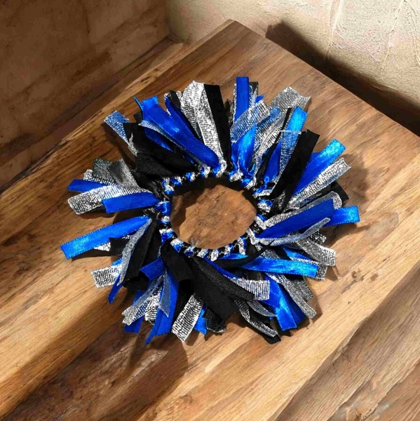 Blue, black, and silver ribbon scrunchie with layered textured strips arranged in a full circular design, displayed on a rustic wooden surface.
