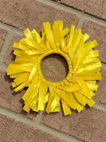 Yellow ribbon fringe scrunchie with shiny satin streamers arranged in a full circular design, photographed on a light brown brick surface background.