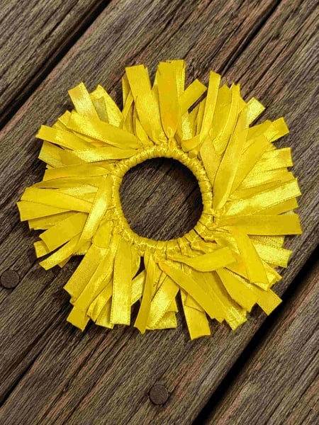 Yellow ribbon fringe scrunchie with layered satin strips arranged in a full circular design, displayed on a rustic weathered wooden deck background.
