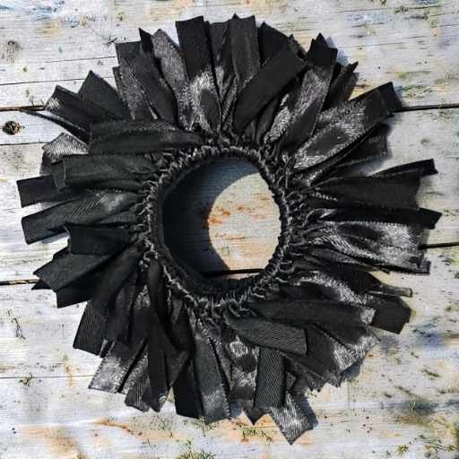 Black ruffled fabric wreath made of layered ribbon strips arranged in a circle on a weathered white wooden surface backdrop for decor.
