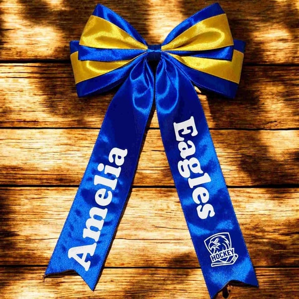 Blue and yellow satin Eagles hockey hair bow with custom Amelia name print, layered ribbon design and glossy finish on rustic wood background.