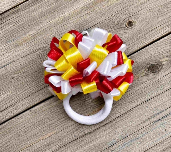 Handmade red, white and yellow ribbon pom pom hair bow on a white elastic hair tie displayed on a rustic wooden surface.