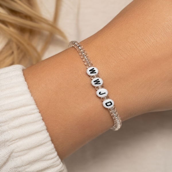 Handmade WWJD seed bead bracelet with clear beads on a stretch band, shown on a woman's wrist against a soft neutral background.