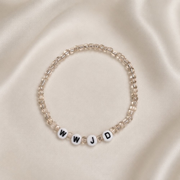 Handmade WWJD clear seed bead stretch bracelet displayed on elegant ivory satin fabric, minimalist Christian faith jewelry gift.