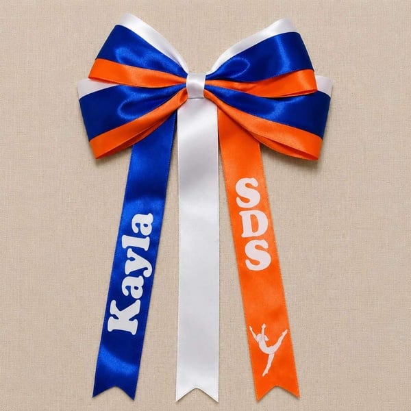 Royal blue, orange, and white layered cheer bow with custom names and SDS on ribbon tails, displayed on neutral linen fabric.