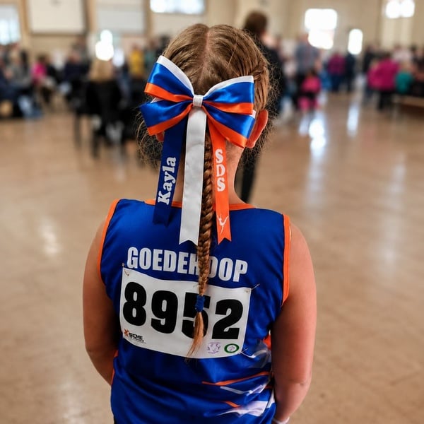 Girl viewed from behind in a blue and orange athletics uniform with a personalized ribbon bow and braid at an indoor event.