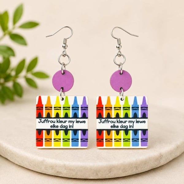 Colorful teacher appreciation dangle earrings with rainbow crayon design and Afrikaans message on a neutral stone display.