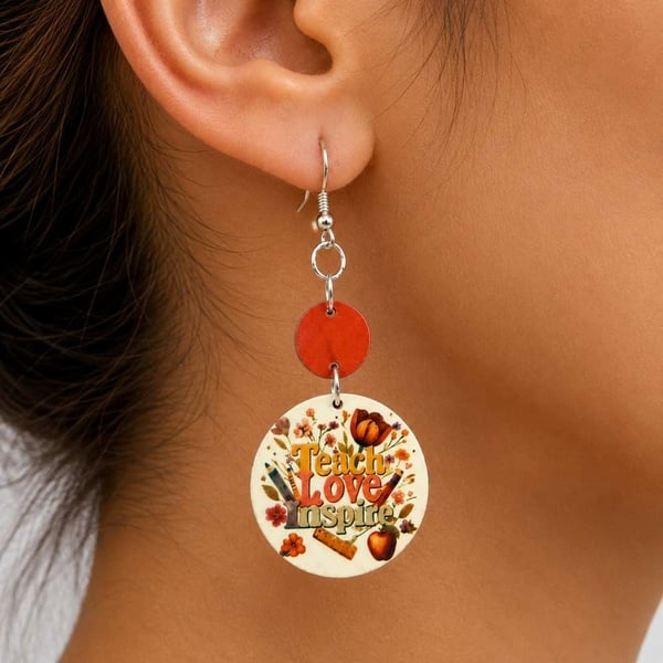 Close-up of woman's ear wearing drop teacher themed earring with red accent and round charm reading Teach Love Inspire with floral design.