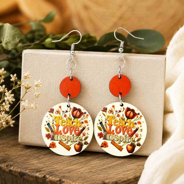 Handmade Teach Love Inspire round MDF drop earrings with orange accents and silver hooks, displayed on a neutral linen stand.