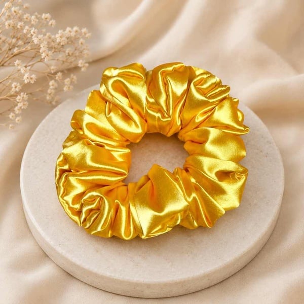 Yellow satin scrunchie showcased on a neutral stone pedestal with soft fabric backdrop, highlighting its silky finish and elegant style.