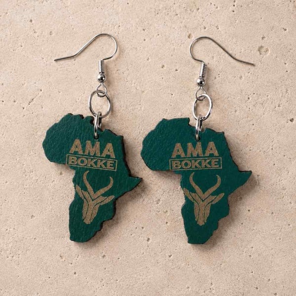 Green Africa-shaped wooden earrings engraved with AMA BOKKE and a Springbok emblem, featuring silver hooks on a neutral stone background.