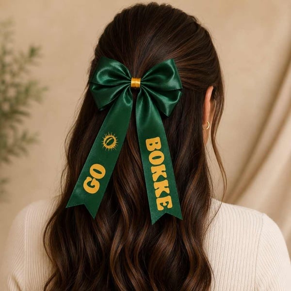 Woman wearing an emerald green Go Bokke satin hair bow with gold print, viewed from behind on a warm neutral background.